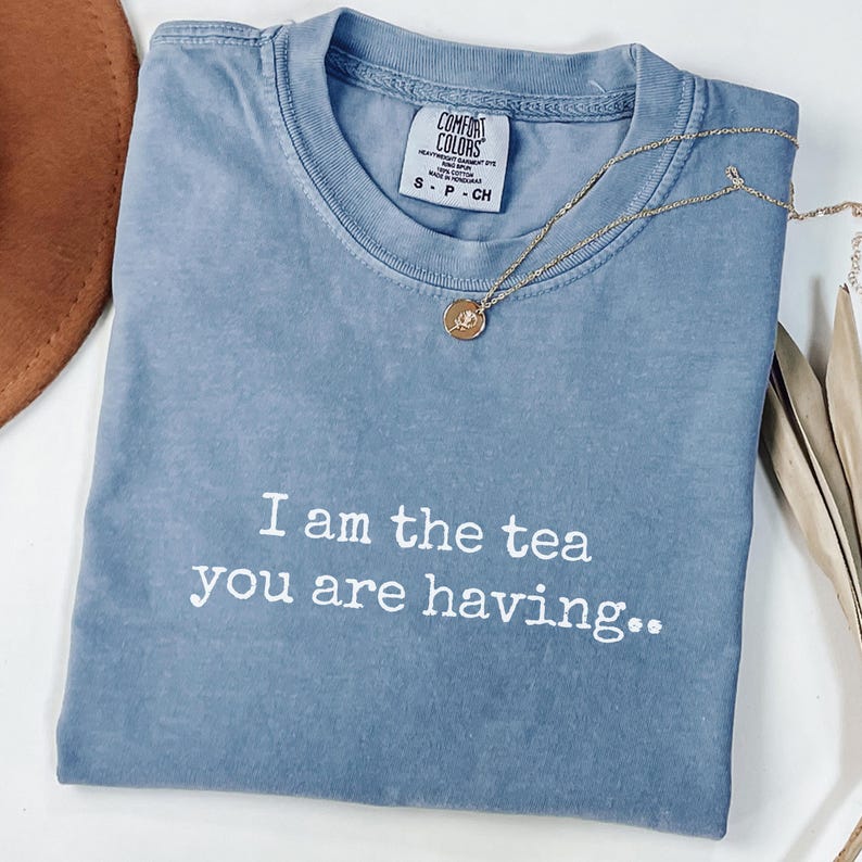 May include: A blue Comfort Colors t-shirt with the text "I am the tea you are having." A gold necklace with a pendant rests on the shirt. The shirt is folded and has a tag at the neckline.