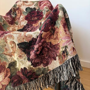 May include: A floral tapestry throw blanket draped over a chair. The blanket features a pattern of large burgundy and pink flowers with green leaves on a beige background. The edges are finished with a black and white fringe.