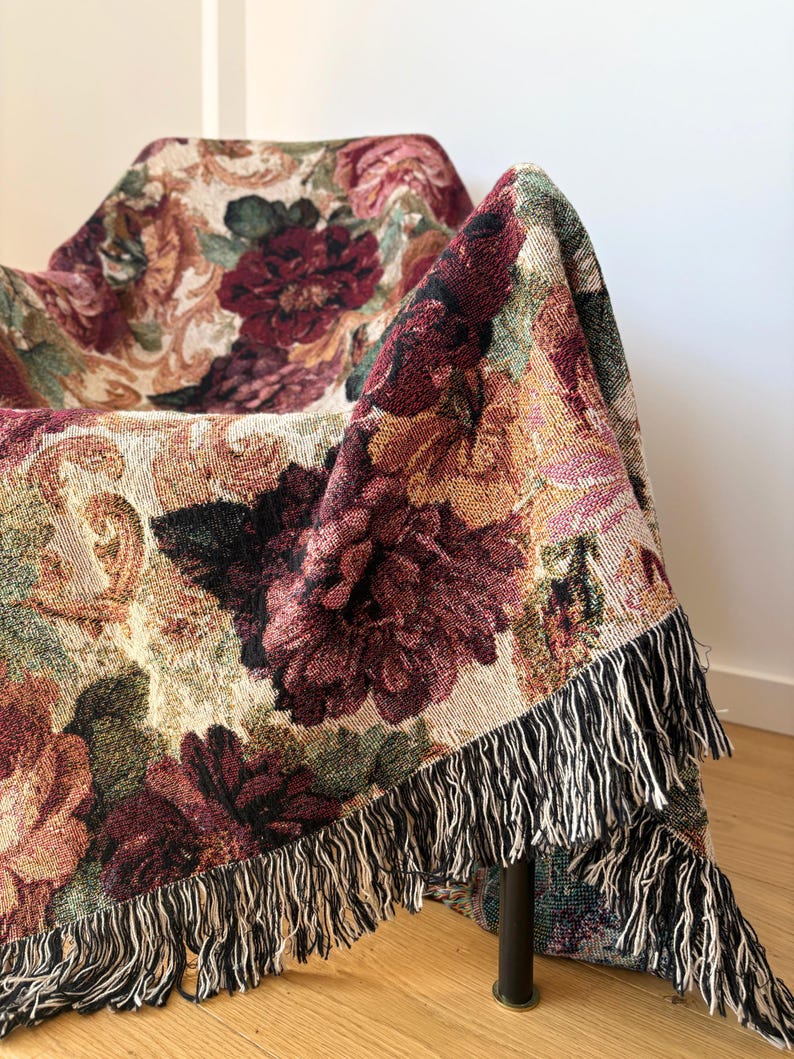 May include: A floral tapestry throw blanket draped over a chair. The blanket features a pattern of large burgundy and pink flowers with green leaves on a beige background. The edges are finished with a black and white fringe.