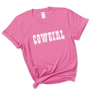 May include: A vibrant pink t-shirt featuring the word "COWGIRL" in white, decorative lettering. The sleeves are rolled up, and the hem is knotted. The Gildan label is visible on the collar.
