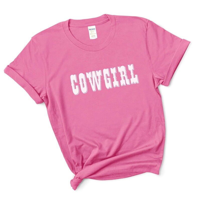 May include: A vibrant pink t-shirt featuring the word "COWGIRL" in white, decorative lettering. The sleeves are rolled up, and the hem is knotted. The Gildan label is visible on the collar.