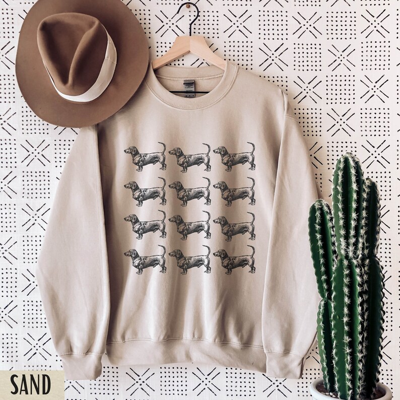 Peut inclure: Un sweat-shirt beige avec un motif r&eacute;p&eacute;t&eacute; de silhouettes de teckels noires. Le sweat-shirt a un col rond et des manches longues.