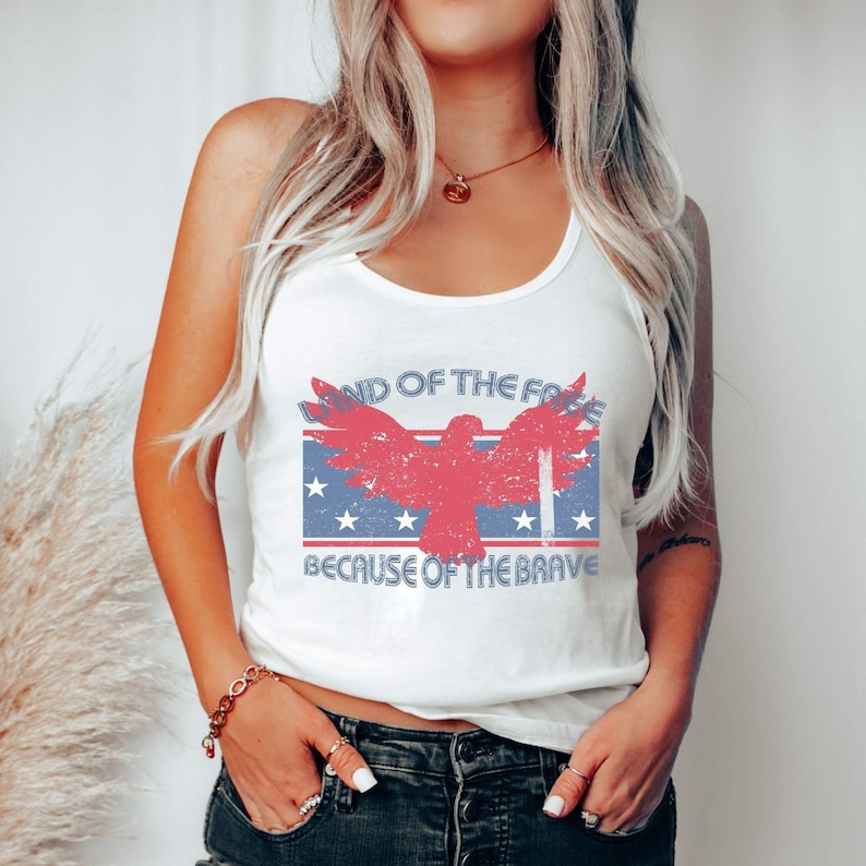May include: White tank top with a red, white, and blue American flag design. The flag features a red eagle with outstretched wings and the text "Land of the Free Because of the Brave".