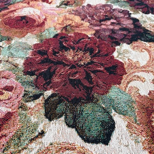 May include: Close-up of a floral tapestry featuring large, detailed roses in shades of burgundy, pink, and peach. The leaves are a mix of green and black. The background has a cream-colored, ornate pattern.