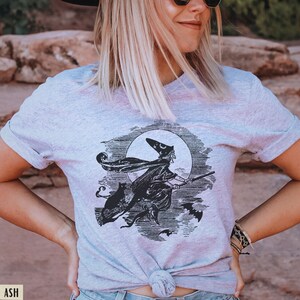 May include: Light gray t-shirt featuring a black and white illustration of a witch flying on a broom in front of a full moon. A black cat and bats are also in the design. The shirt is tied at the waist.