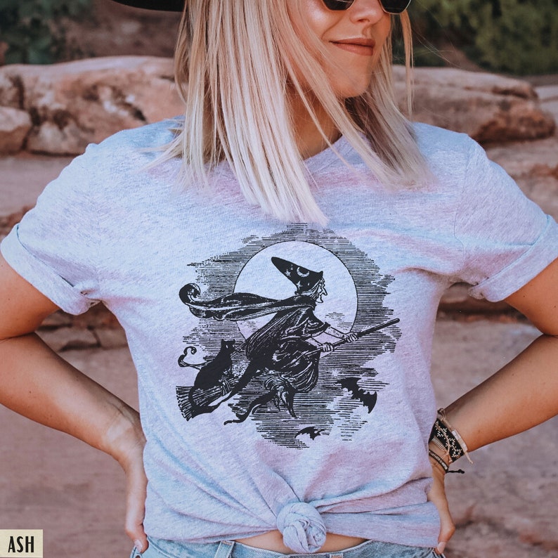 May include: Light gray t-shirt featuring a black and white illustration of a witch flying on a broom in front of a full moon. A black cat and bats are also in the design. The shirt is tied at the waist.