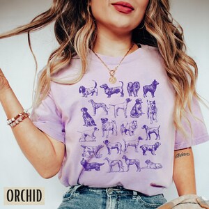 May include: A purple t-shirt with a graphic print of various dog breeds in a vintage style. The shirt has a crew neck and short sleeves.