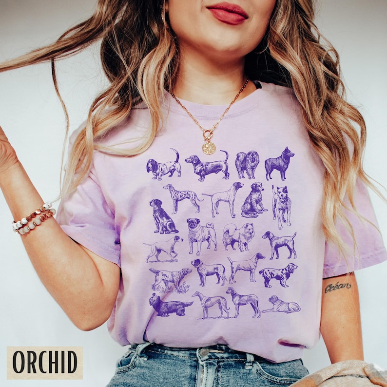 May include: A purple t-shirt with a graphic print of various dog breeds in a vintage style. The shirt has a crew neck and short sleeves.