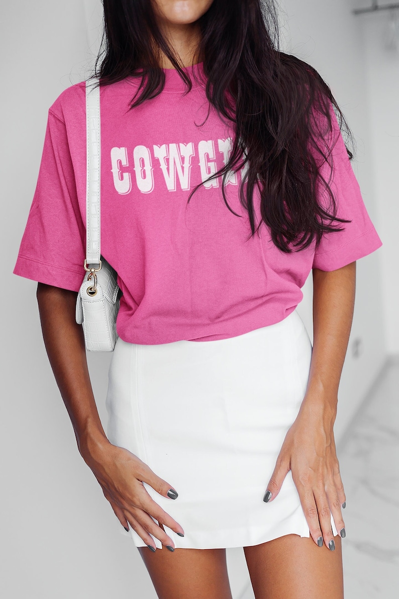 May include: A magenta t-shirt with the word "COWGIRL" in white lettering, worn with a white skirt and a white crossbody bag. The outfit is completed with dark nail polish.