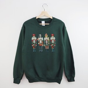 May include: A dark green sweatshirt featuring a watercolor illustration of four toy nutcracker soldiers. The nutcrackers are in various colors, including red, gold, and blue, and are standing in a row. The sweatshirt is hanging on a wooden hanger.