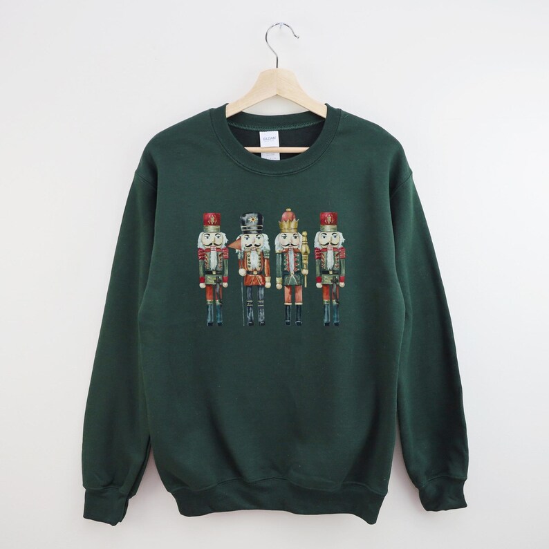 May include: A dark green sweatshirt featuring a watercolor illustration of four toy nutcracker soldiers. The nutcrackers are in various colors, including red, gold, and blue, and are standing in a row. The sweatshirt is hanging on a wooden hanger.