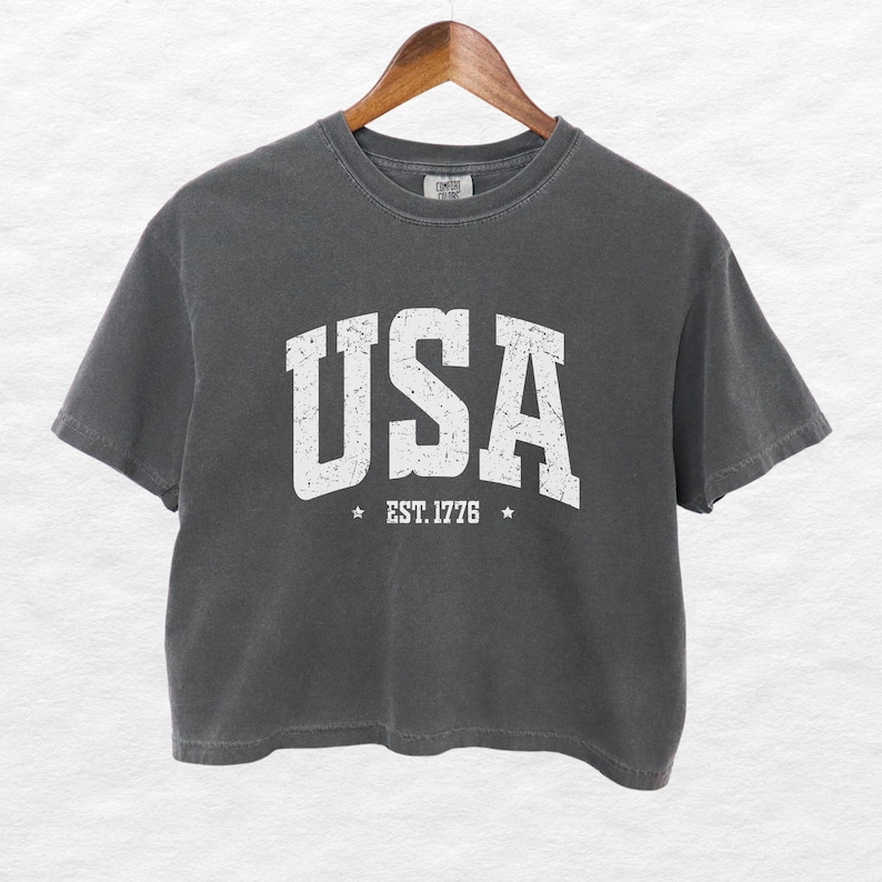 Pu&ograve; includere: Una maglietta corta grigio antracite con la scritta "USA" in lettere bianche invecchiate. Sotto la parola "USA" c'&egrave; il testo "EST. 1776" con una stella su ogni lato. La maglietta &egrave; appesa a una gruccia di legno.