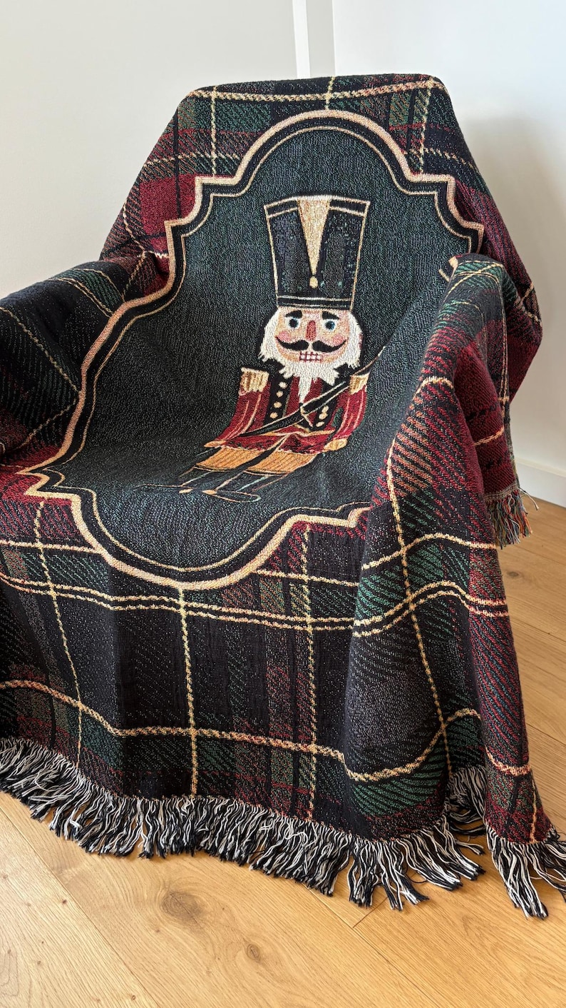 May include: A woven throw blanket draped over a chair, featuring a plaid pattern in black, green, and red. The center showcases a detailed illustration of a nutcracker soldier in a decorative frame. The blanket has a fringed edge.