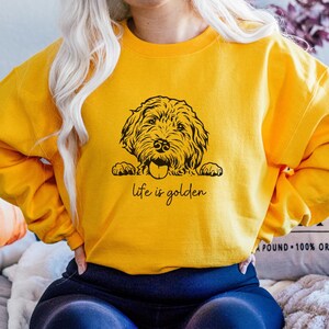 以下が含まれることがあります： 黄色いスウェットシャツに、ゴールデン・ドゥードルの犬が上の方から顔を出している黒いイラストが描かれています。犬の下には「life is golden」という文字が印刷されています。