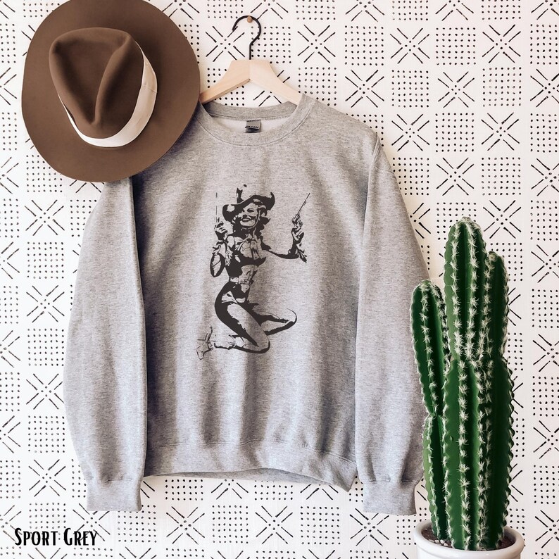 Pu&ograve; includere: Una felpa grigia con una silhouette nera di una donna che indossa un cappello da cowboy e tiene due pistole. Il testo "SPORT GREY" &egrave; visibile nella parte inferiore dell'immagine.
