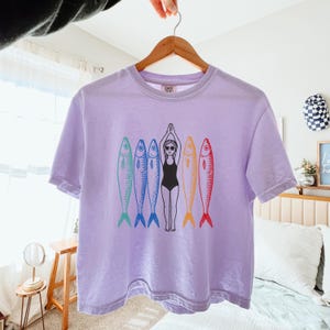 以下が含まれることがあります： 紫色のTシャツに、水着を着た女性が5匹のカラフルな魚に囲まれているグラフィックがプリントされています。魚はすべて異なる色で、黒いアウトラインがあります。