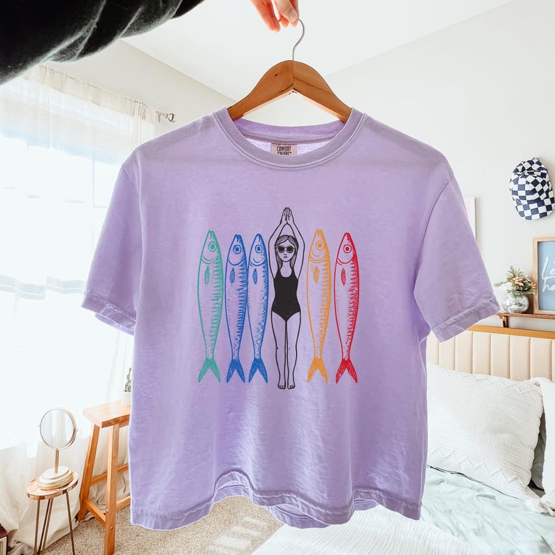 以下が含まれることがあります： 紫色のTシャツに、水着を着た女性が5匹のカラフルな魚に囲まれているグラフィックがプリントされています。魚はすべて異なる色で、黒いアウトラインがあります。
