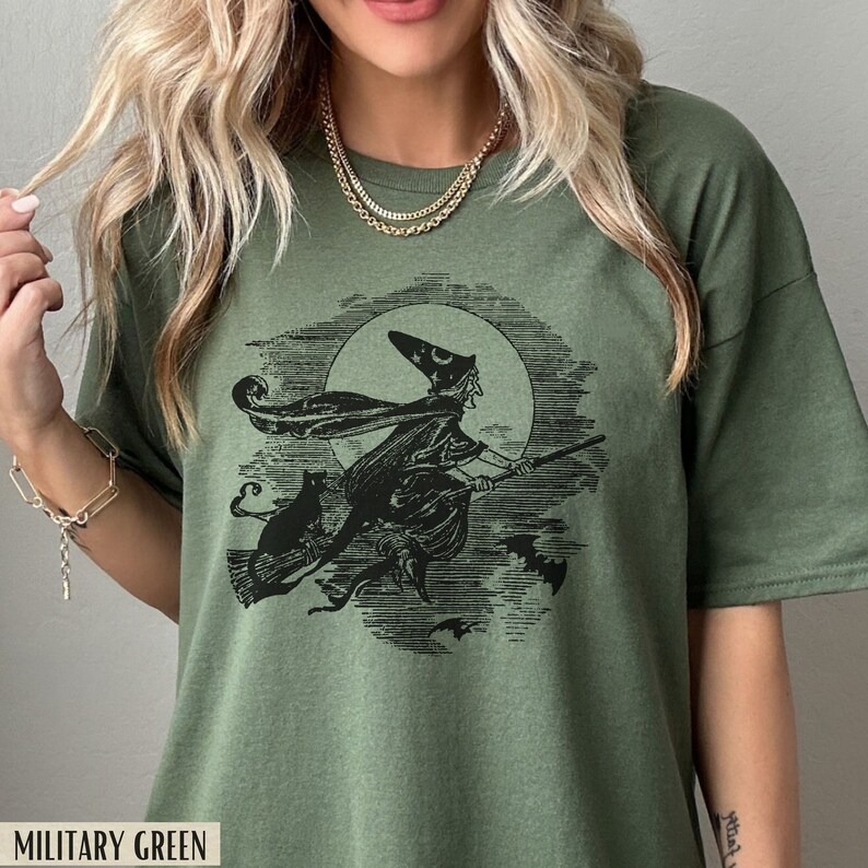 May include: Military green t-shirt featuring a black and white illustration of a witch on a broomstick, with a black cat and bats. The witch is flying in front of a full moon. The text "MILITARY GREEN" is at the bottom.