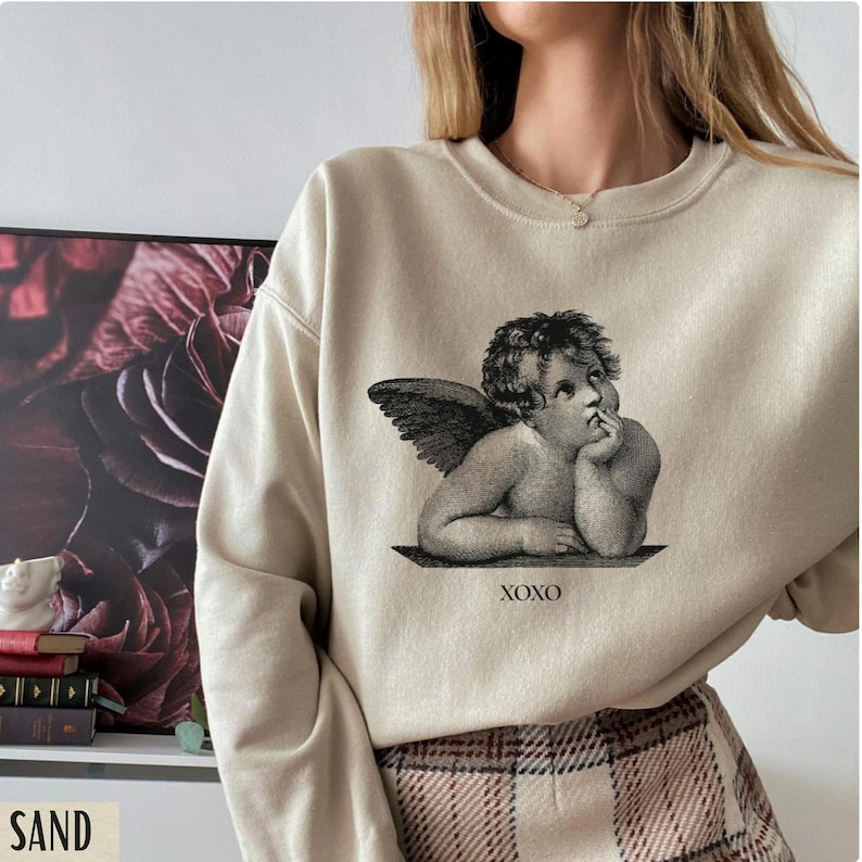 Op de afbeelding: Een beige sweatshirt met een zwart-witte grafische print van een cherubijn met vleugels. De grafiek bevat de tekst "XOXO".