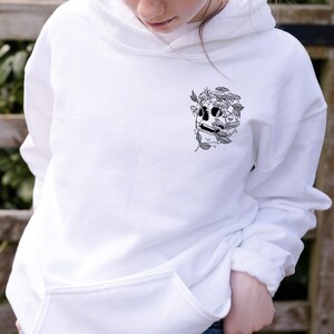 May include: White hoodie with a black and white skull graphic on the chest. The skull is decorated with mushrooms and leaves. The hoodie has a front pocket and a hood.