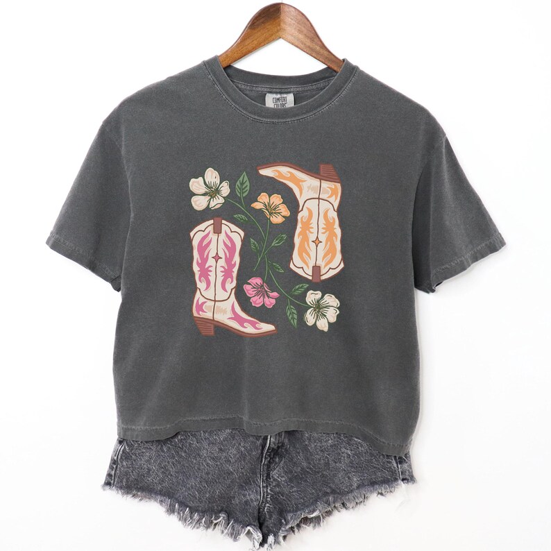 May include: Dark grey t-shirt with a graphic of two cowboy boots in pink and orange, and floral accents. The shirt is paired with grey denim shorts. The design is centered on the front of the shirt.