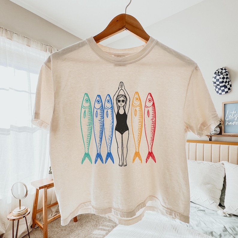 以下が含まれることがあります： クリーム色のクロップドTシャツに、黒い水着を着た女性が5匹のカラフルな魚と一緒に泳いでいるグラフィックがプリントされています。魚は青、緑、黄、オレンジ、赤です。