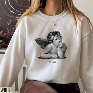 Op de afbeelding: Witte trui met een zwart-witte grafische print van een cherubijn met vleugels en de tekst "XOXO".