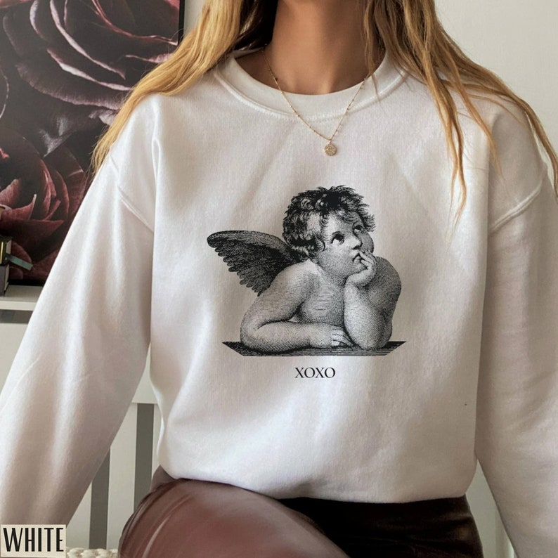 Op de afbeelding: Witte trui met een zwart-witte grafische print van een cherubijn met vleugels en de tekst "XOXO".