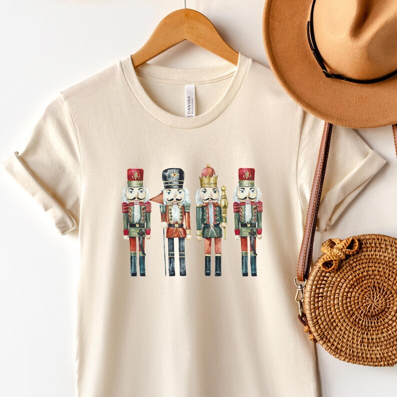 May include: Cream-colored t-shirt featuring a watercolor illustration of four toy nutcrackers. The nutcrackers are dressed in red, green, and gold uniforms with black boots. The shirt is hanging on a wooden hanger.