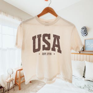 Pu&ograve; includere: Top corto color crema con la scritta "USA" in lettere marroni effetto invecchiato. Sotto, le parole "EST. 1776" con una stella su ogni lato. La maglietta &egrave; appesa a una gruccia di legno.