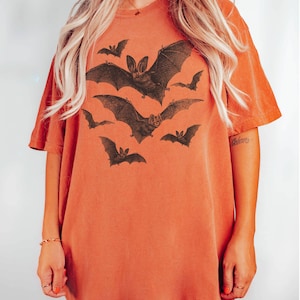 May include: A burnt orange oversized t-shirt with a graphic print of bats in flight. The bats are black and white and have a vintage illustration style.