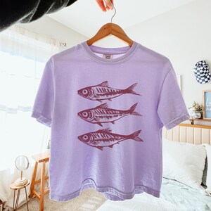 Puede incluir: Una camiseta de color lavanda con un corte corto presenta un gr&aacute;fico de tres peces estilizados en color marr&oacute;n. La camiseta tiene mangas cortas y cuello redondo.