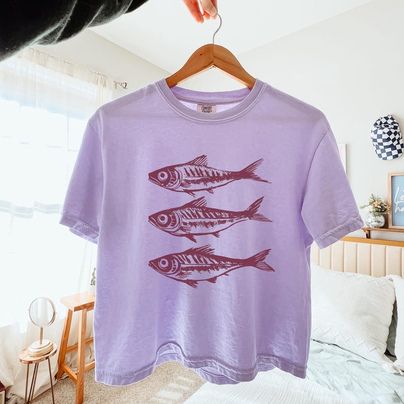 Puede incluir: Una camiseta de color lavanda con un corte corto presenta un gr&aacute;fico de tres peces estilizados en color marr&oacute;n. La camiseta tiene mangas cortas y cuello redondo.