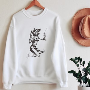 Pu&ograve; includere: Felpa bianca con una stampa grafica in bianco e nero di una donna che indossa un cappello da cowboy e tiene due pistole.