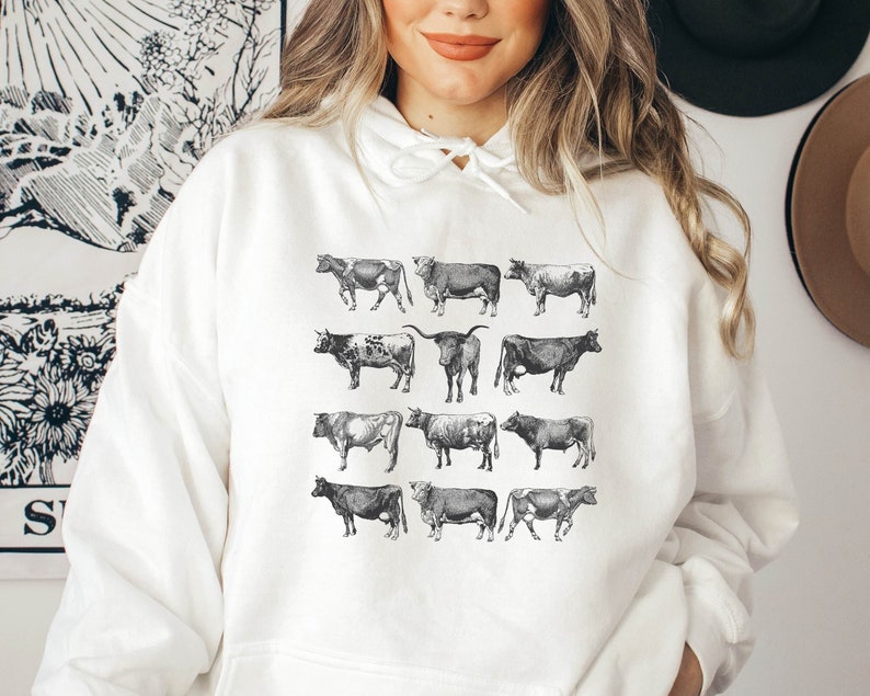 May include: White hoodie with a black and white illustration of cows on the front. The illustration features 12 cows in various poses.
