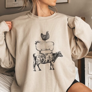 May include: A beige sweatshirt with a graphic print of a cow, sheep, and chicken stacked on top of each other. The animals are black and white.