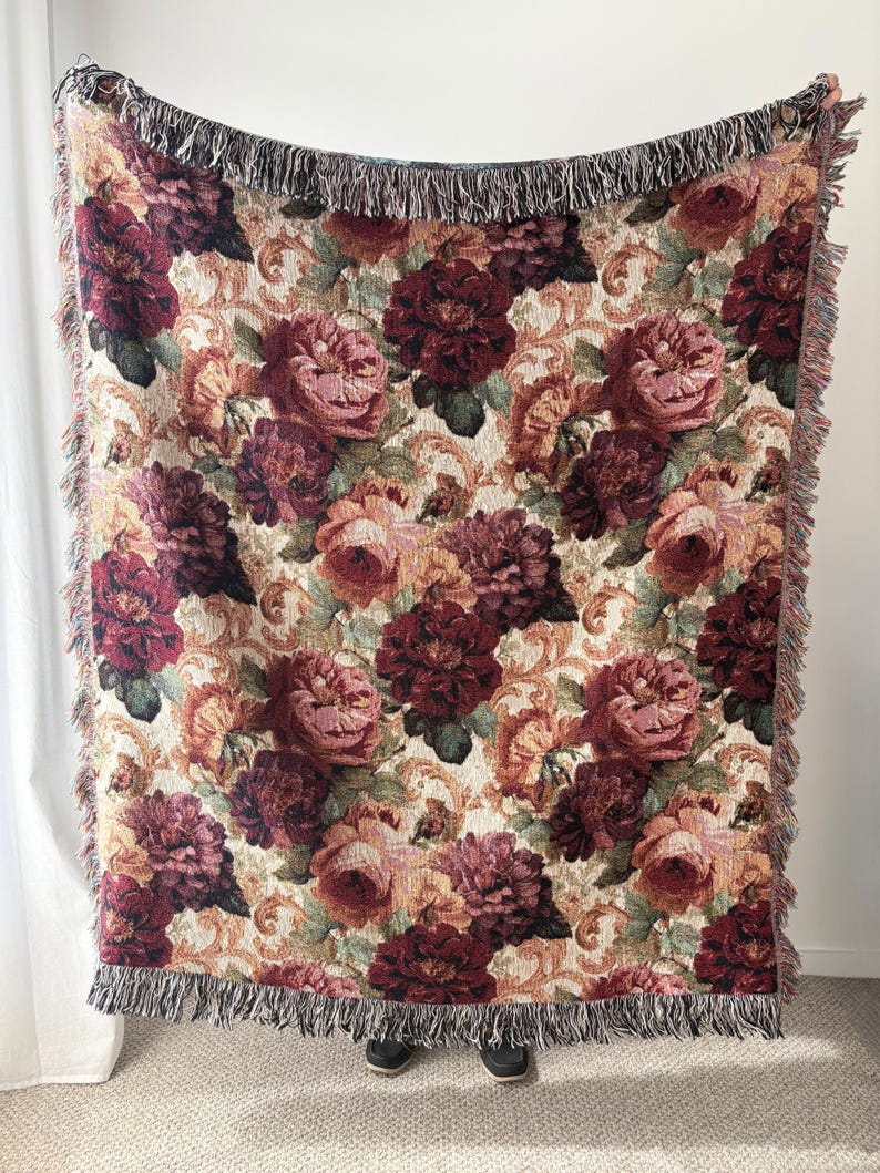 May include: Woven throw blanket with a floral pattern featuring large burgundy and pink roses on a cream background. The blanket has a fringed edge in shades of brown and black. The overall design has a vintage aesthetic.