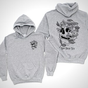 May include: Two light gray hoodies with a black graphic of a skull with mushrooms and leaves. The back of one hoodie has the text "Not Dead Yet" below the skull design. The other hoodie has the same skull design on the front.