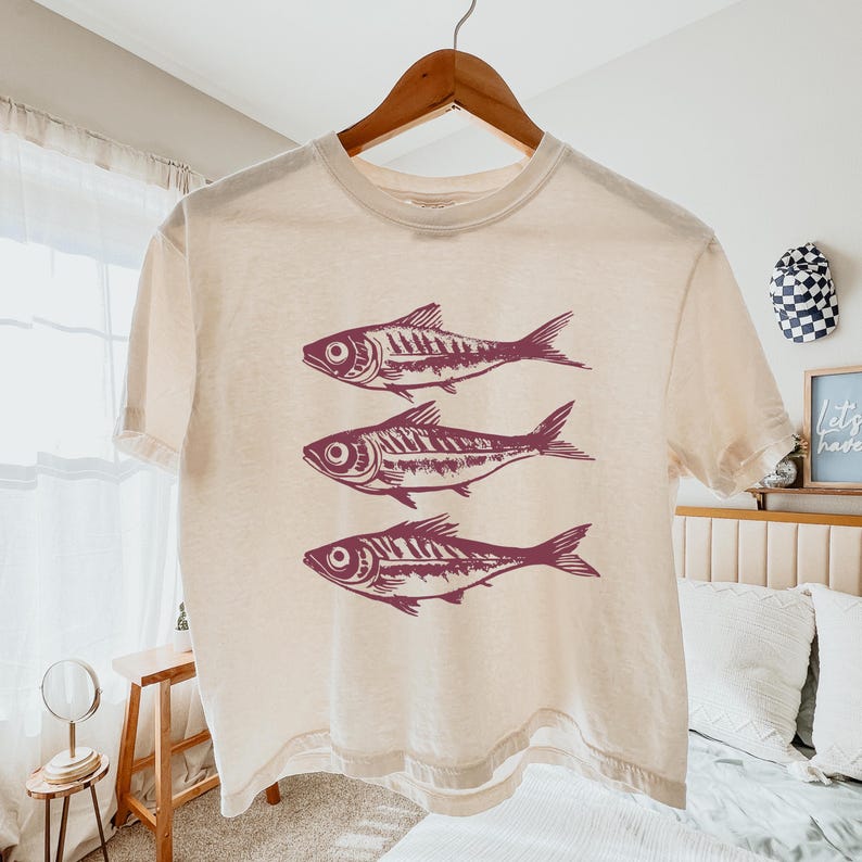 Puede incluir: Una camiseta recortada de color crema con un gr&aacute;fico de tres peces rojos en la parte delantera.