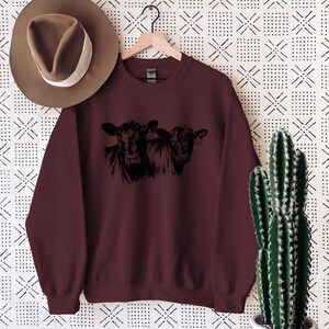 K&ouml;nnte beinhalten: Burgunderfarbenes Sweatshirt mit einer schwarzen Grafik von zwei Kuhgesichtern. Das Sweatshirt h&auml;ngt an einem schwarzen Kleiderb&uuml;gel vor einem gemusterten Hintergrund. Auf der linken Seite des Bildes ist ein brauner Hut zu sehen.