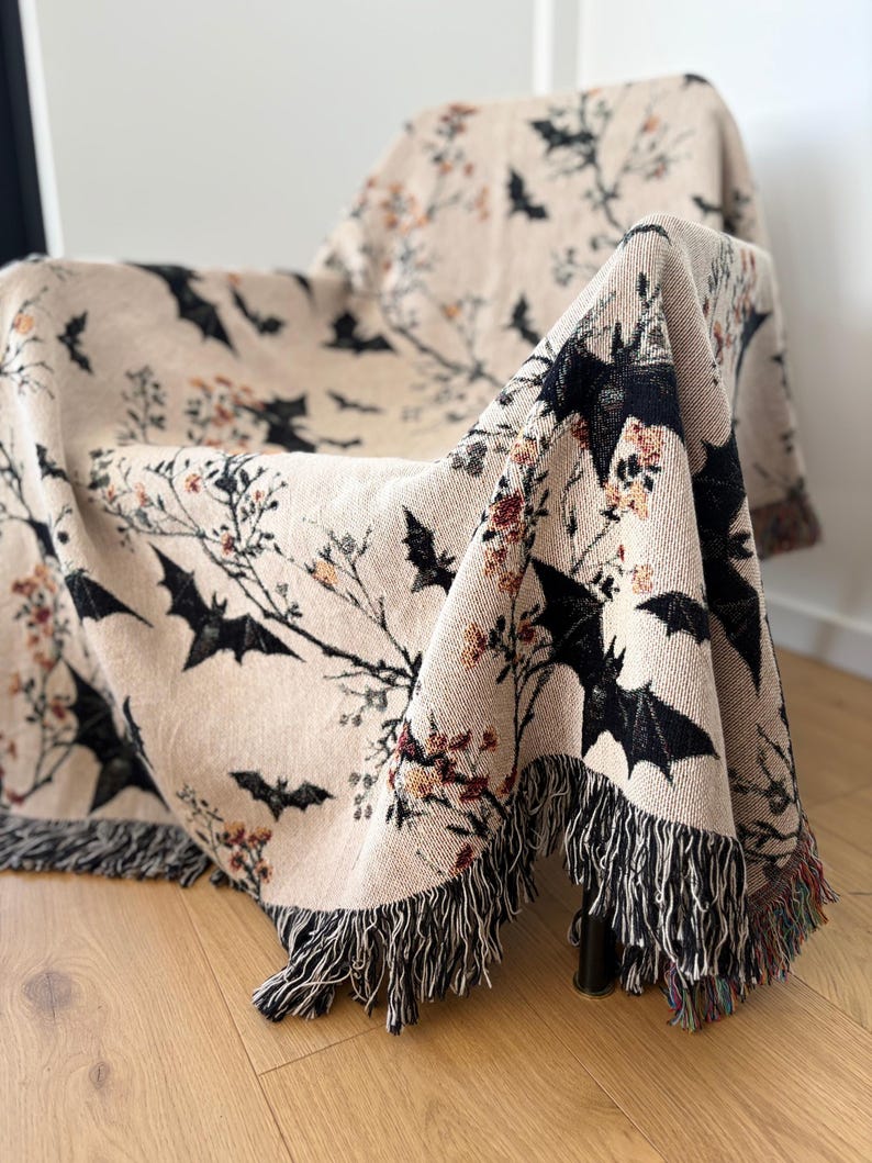 May include: A beige throw blanket draped over a chair, featuring a repeating pattern of black bats and floral branches. The blanket has a fringed edge with black and multicolored threads, adding texture and detail to the Halloween-themed home decor.