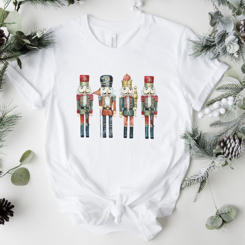 May include: White t-shirt featuring a watercolor illustration of four toy nutcracker soldiers. The nutcrackers are dressed in red, green, and gold uniforms with black boots. The shirt is a crew neck and has a knot at the bottom.