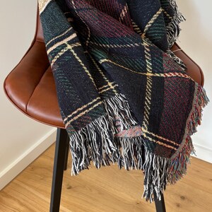 May include: A woven throw blanket with a plaid pattern in black, green, red, and gold, draped over a brown leather chair. The blanket has a fringe of white and multicolored threads along the edges. The chair has a black metal frame.