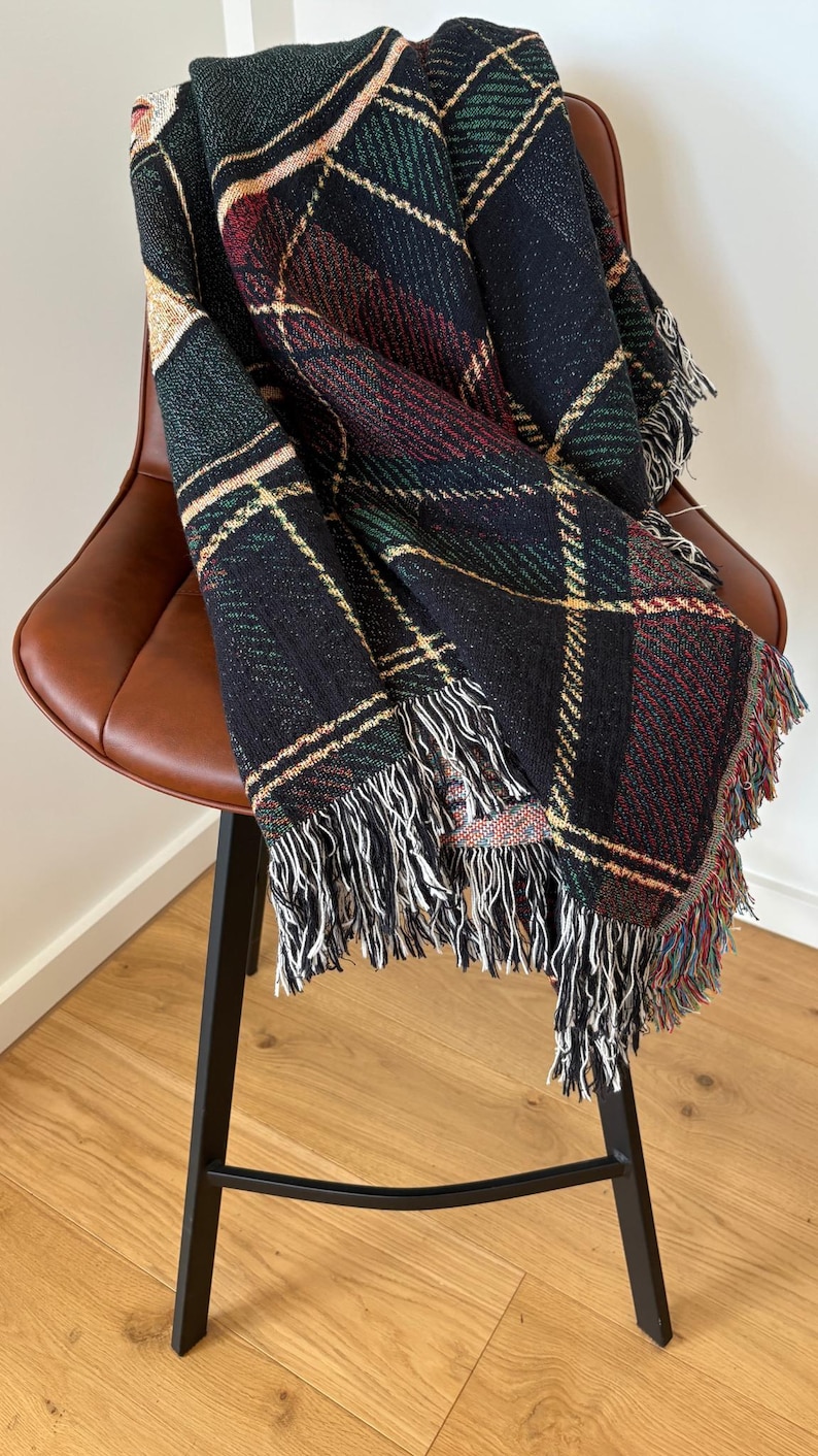 May include: A woven throw blanket with a plaid pattern in black, green, red, and gold, draped over a brown leather chair. The blanket has a fringe of white and multicolored threads along the edges. The chair has a black metal frame.