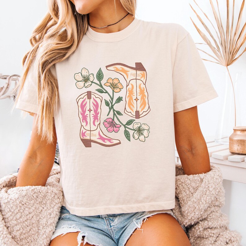 May include: Cream-coloured t-shirt with a graphic of cowboy boots and flowers in pink, orange, and green. The boots have pink and orange detailing. The design is centred on the shirt.