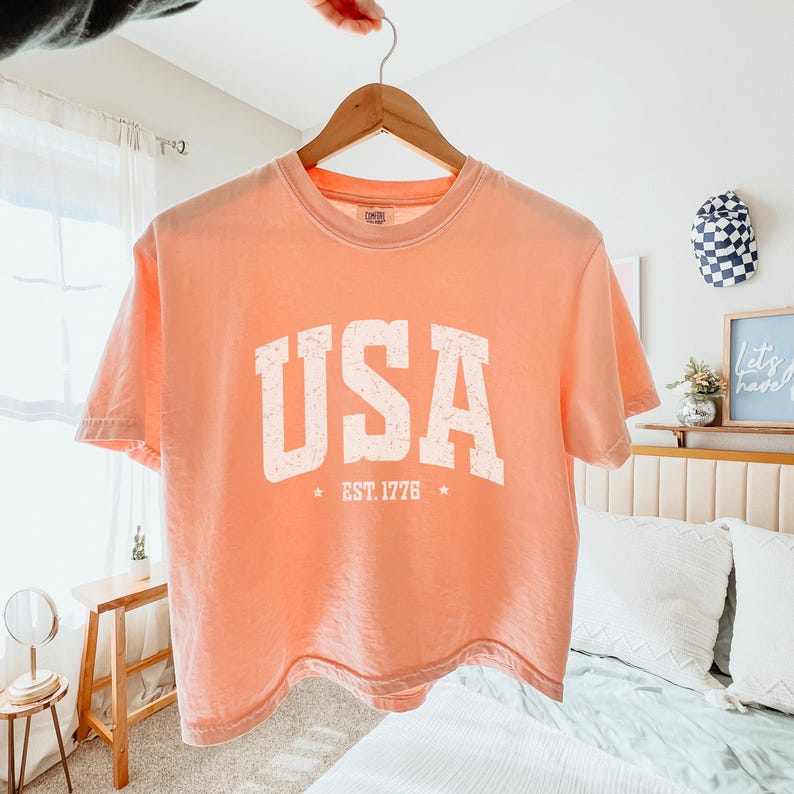 Pu&ograve; includere: T-shirt color pesca con la scritta "USA" in lettere bianche invecchiate. Sotto, "EST. 1776" con stelle su entrambi i lati. La maglietta &egrave; appesa a una gruccia di legno.
