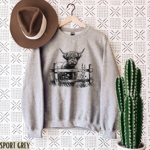 Puede incluir: Una sudadera gris con una ilustraci&oacute;n en blanco y negro de una vaca de las Tierras Altas de Escocia de pie en un campo de hierba y flores. El texto "Sport Grey" es visible en la parte inferior de la imagen.