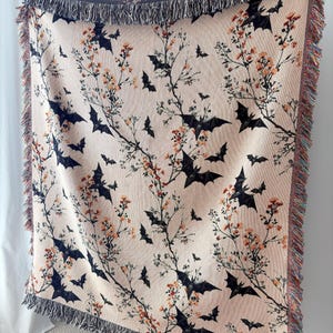 May include: A beige woven throw blanket featuring a pattern of black bats and flowering branches. The blanket has a fringed edge in shades of gray, orange, and red. The design is suitable for Halloween or autumn decor.