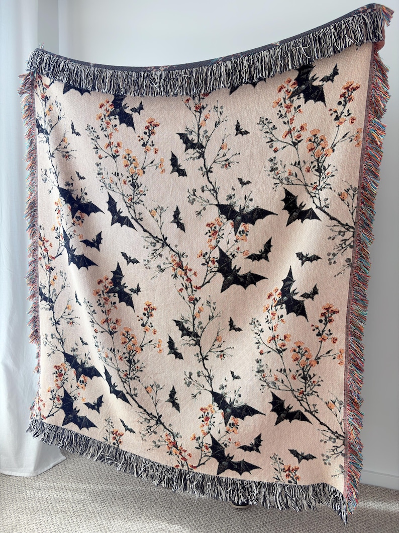 May include: A beige woven throw blanket featuring a pattern of black bats and flowering branches. The blanket has a fringed edge in shades of gray, orange, and red. The design is suitable for Halloween or autumn decor.