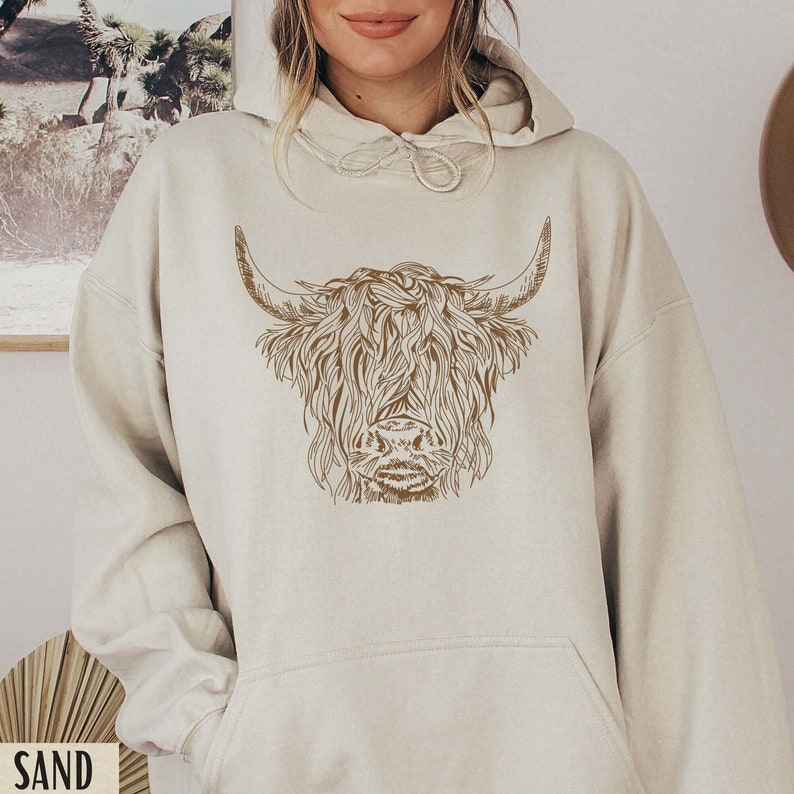 May include: A cream-colored hoodie with a brown outline of a Highland cow's head on the front.